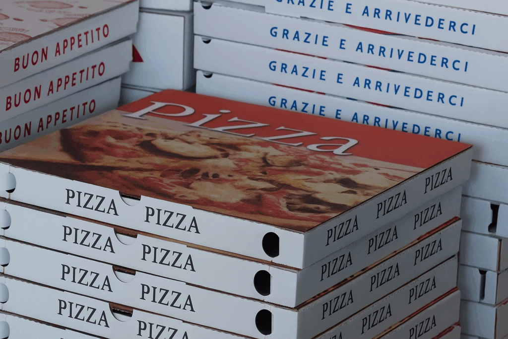 Stack di scatole da pizza bianche con scritte “Pizza”, “Buon appetito” e “Grazie e arrivederci” impilate in un ambiente di pizzeria, riprese in primo piano.